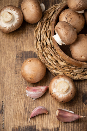 Aerial view of portobello mushrooms in wooden bowl on wooden table with more mushrooms and garlic, vertical