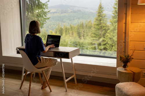 Woman works on laptop while sitting by the table in front of panoramic window with great view on mountains. Remote work and escaping to nature concept