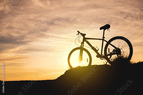 Fototapeta Naklejka Na Ścianę i Meble -  silhouette of a bicycle on a hill against the sky during sunset. evening bike ride, the bike is in the grass