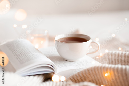 Open book with cup of coffee in bed with knit cloth sweater over glow christmas lights at background close up. Good morning.
