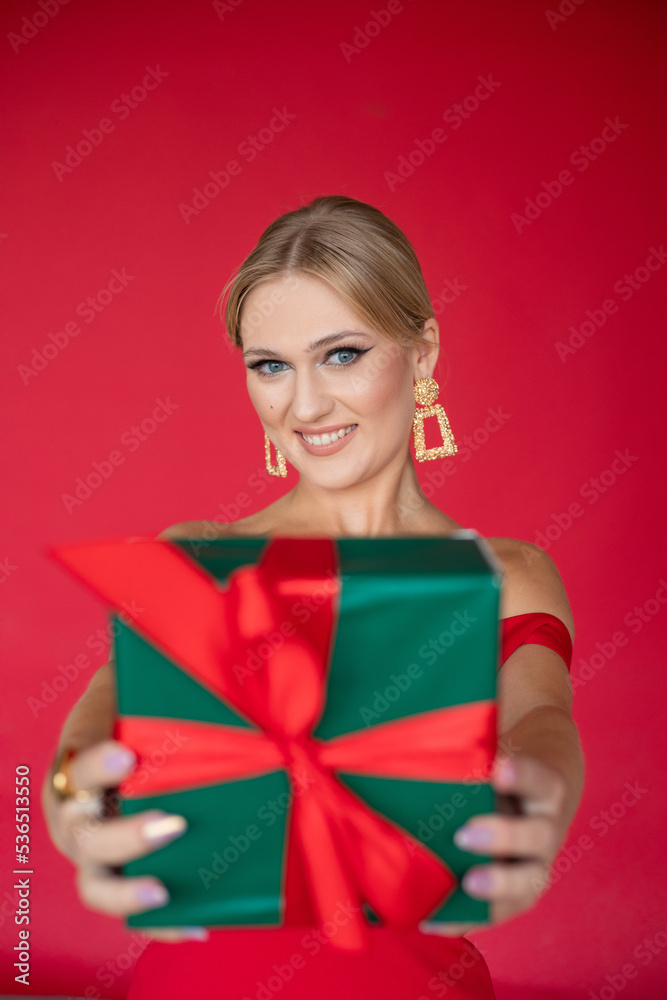Vertical smiling elegant woman in gala dress offering, showing green present box with ribbon on red studio background. Vertical smiling elegant woman in gala dress offering, showing green present box with ribbon on red studio background.