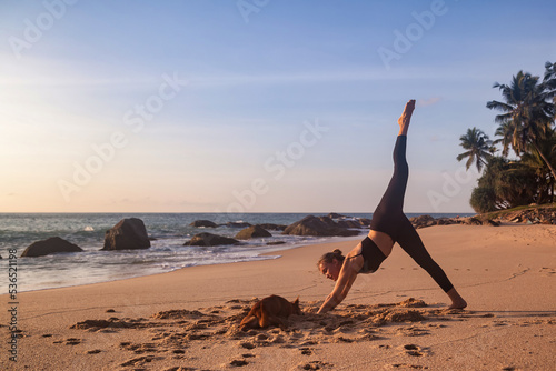 Young woman with doggy does asana yoga for healthy lifestyle on tropical ocean beach outdoors. Female performing sports exercises to restore strength and spirit. Yoga position on seashore. Copy space