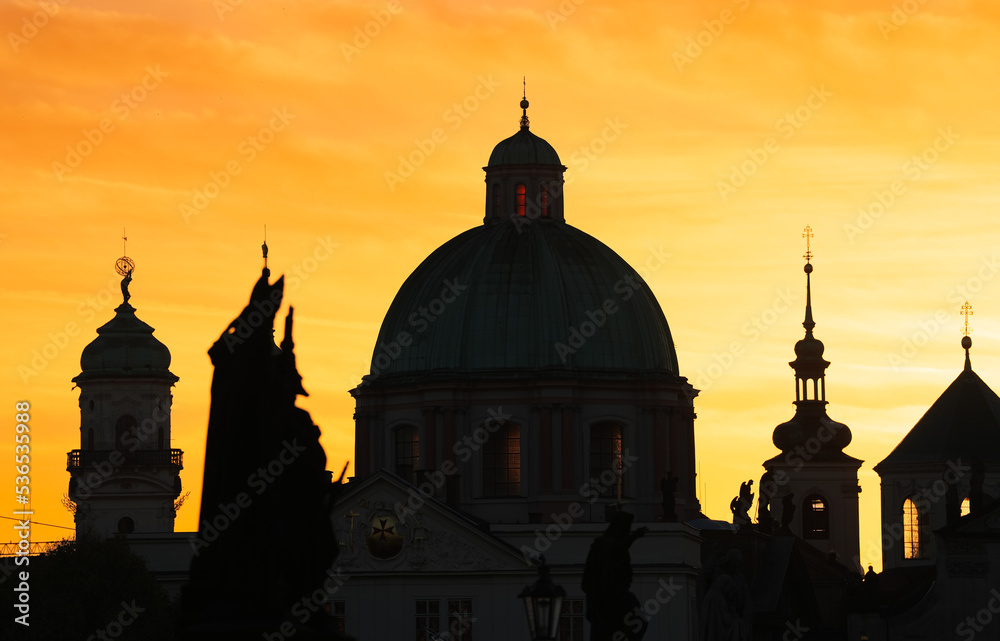 Silhouettes of the metalic Christian Orthodox and Catholic cross on top of a cathedral from Prague during spectacular sunrise with orange sky.