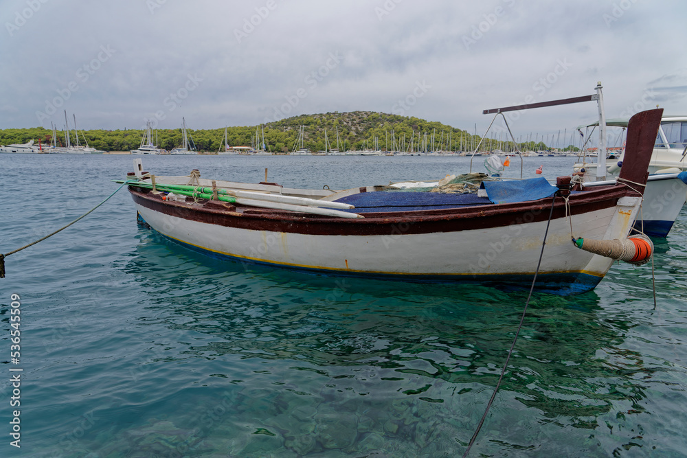 White fishing boat for rowing in the beautiful blue crystal clear waters of the Croatian Mediterranean. In the background there is a marina and green mountains.