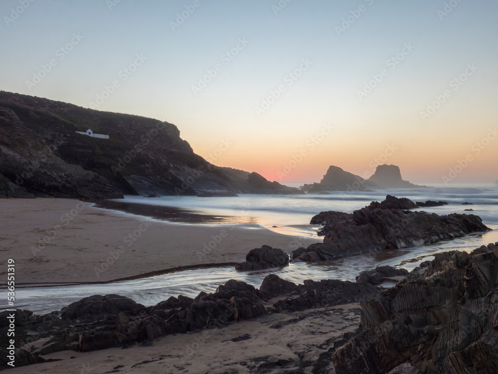 After sunset blue hour long exposure view of sand beach Praia da Zambujeira do Mar with rock and cliff, small stream and with blurred ocean waves in pink light. Rota Vicentina coast, Odemira, Portugal