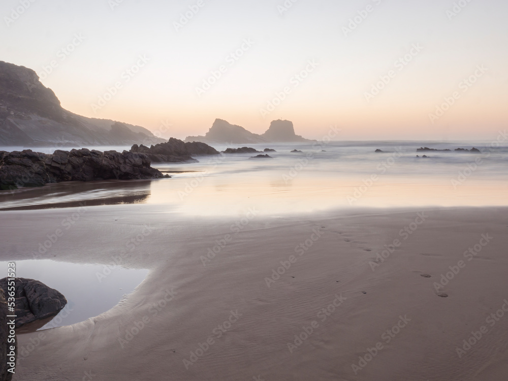 After sunset blue hour dreamy long exposure view of sand beach Praia da Zambujeira do Mar with rock and cliff and blurred ocean waves in pink light. Rota Vicentina coast, Odemira, Portugal