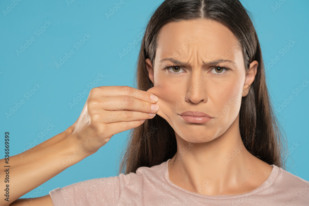 Portrait of young unhappy brunette woman, pulling her cheek skin on blue background Portrait of young unhappy brunette woman, pulling her cheek skin on blue background