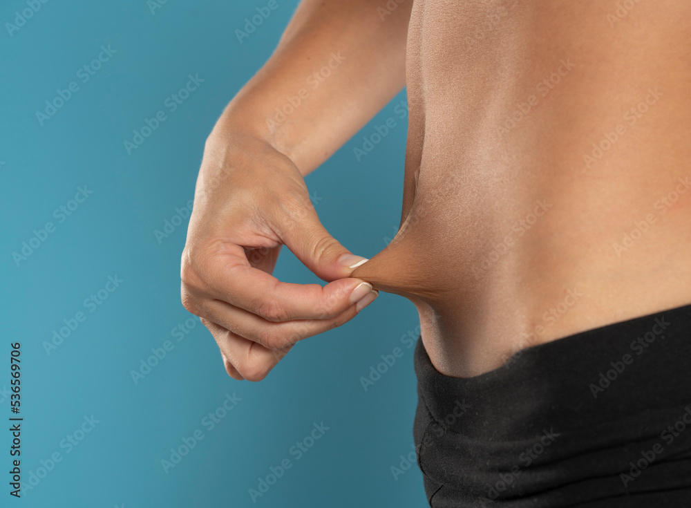 Closeup of a young fit woman pinching her belly skin on a blue background Closeup of a young fit woman pinching her belly skin on a blue background