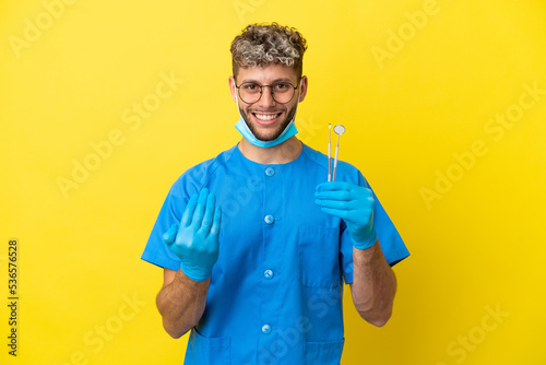 Dentist caucasian man holding tools isolated on yellow background inviting to come with hand. Happy that you came