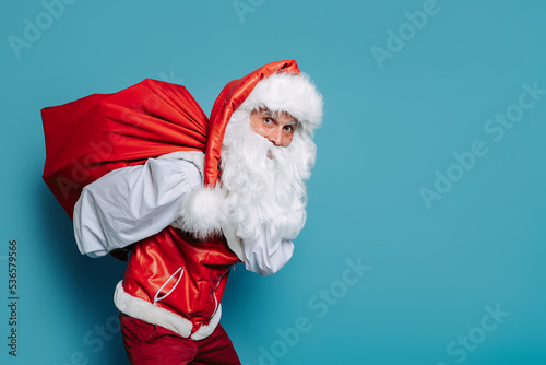 slinking santa claus carrying huge bag of gifts on blue background while looking at camera.