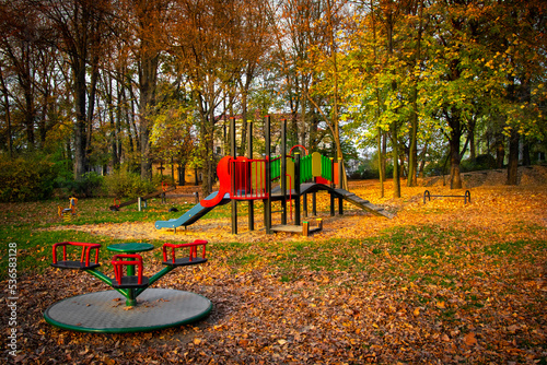 Trees with colorful autumn foliage. Children's empty playground with slides and swings is covered with yellow maple leaves.  photo