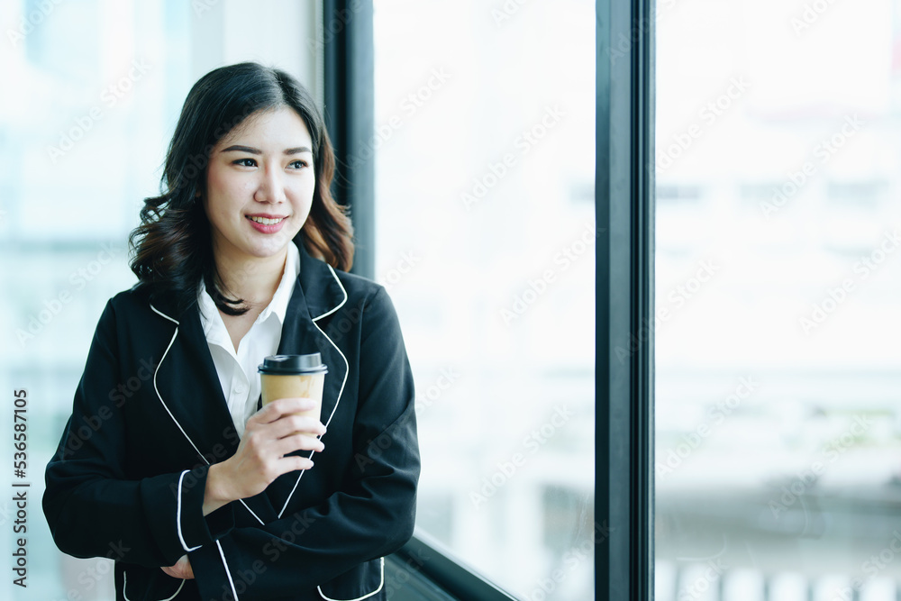 A portrait of a young Asian businesswoman smiling happily and drinking coffee by the window in the conference room