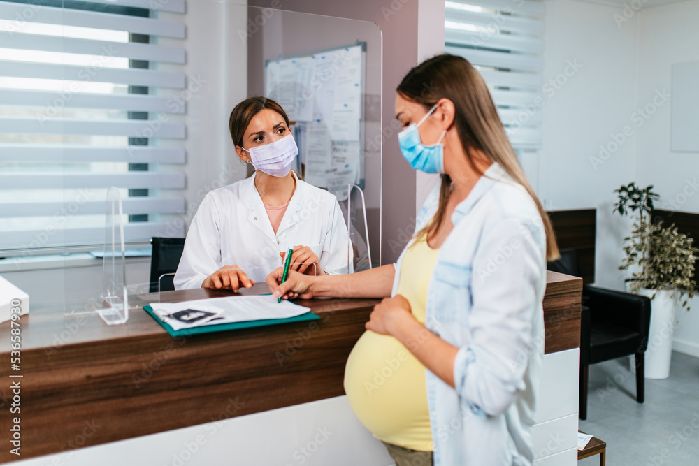 The pregnant woman is scheduling the next check-up with a gynecologist at the hospital. The nurse and pregnant woman are wearing protective face masks due to the coronavirus pandemic. The pregnant woman is scheduling the next check-up with a gynecologist at the hospital. The nurse and pregnant woman are wearing protective face masks due to the coronavirus pandemic.