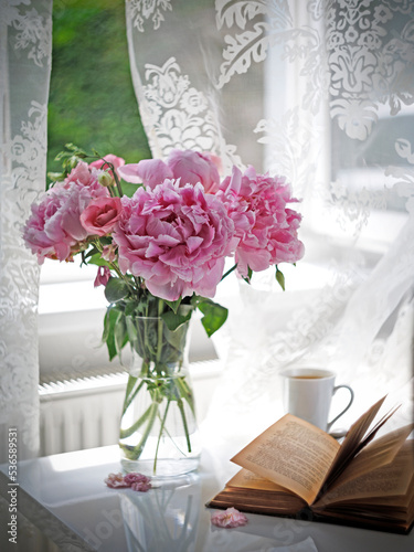 Bouquet of chic pink peonies in a transparent glass vase and an open book on the windowsill photo