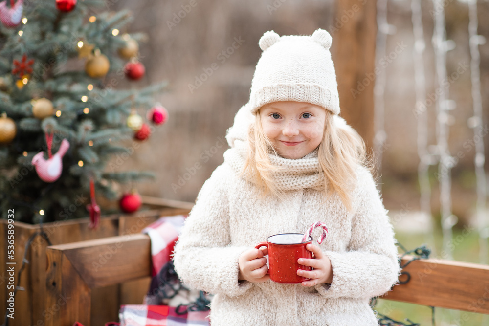 Cute smiling kid girl 4-5 year old drink hot chocolate with red candy stick in mug over Chrismas tree decoration outdoor close up. Happy winter holidays. Cute smiling kid girl 4-5 year old drink hot chocolate with red candy stick in mug over Chrismas tree decoration outdoor close up. Happy winter holidays.