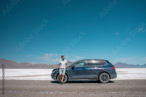 Man standing by car at Bonneville salt flat. Male tourist is relaxing at tranquil landscape with clear sky in background. He is spending leisure time at famous place during summer. © Aerial Film Studio