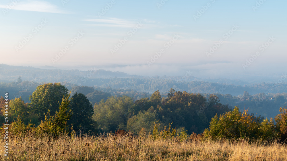 Atmospheric hilly rural landscape with morning fog in autumn, hills overgrown with forest