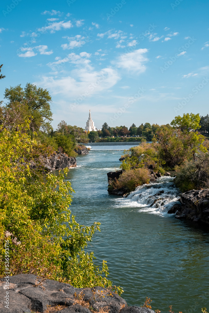 View of famous temple seen from beautiful Idaho falls. Snake river flowing through rocks and plants with sky in background. Picturesque scenery of scenic waterfalls and tourist attraction in city. View of famous temple seen from beautiful Idaho falls. Snake river flowing through rocks and plants with sky in background. Picturesque scenery of scenic waterfalls and tourist attraction in city.