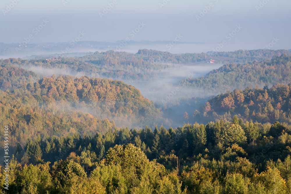 Morning fog in valleys between hills in autumn, airy rural landscape with deciduous forest