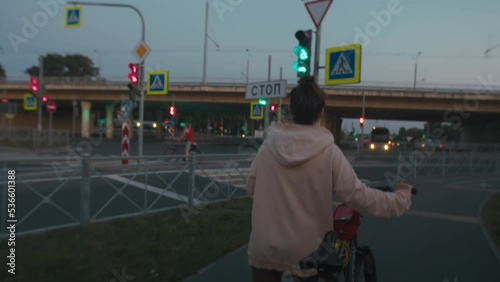 Hanheld shot of young woman in casual sport clothes walks along the street by night and riding her bike to crosswalk. Female come back home after her daily routine evening stroll, city nightlife with