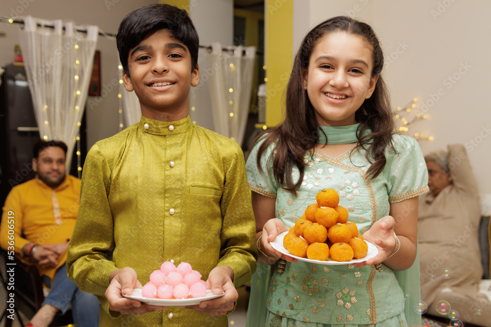 Young girl and boy, kid, dressed up in ethnic wear holding plate of sweets with smile expression pose for photo with family in background celebrating diwali Hindu festival Laxmi poojan