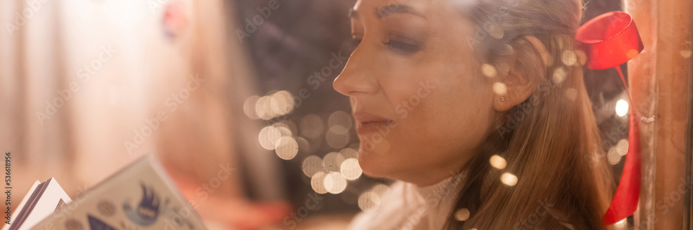 blurry portrait of a candid smiling woman face through the window glass enjoying reading a hygge book and relaxing on happy new year and merry christmas eve. bokeh of glowing lights garland. banner blurry portrait of a candid smiling woman face through the window glass enjoying reading a hygge book and relaxing on happy new year and merry christmas eve. bokeh of glowing lights garland. banner