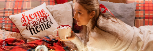 candid young woman with cup of cocoa drink and marshmallows and smartphone at home in a cozy bed on a red blanket or plaid enjoyment happy new year and merry christmas eve. comfy slow living. banner