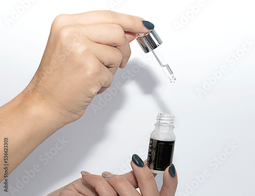 Female hands hold a small bottle with liquid and a pipette with a drop falling on a white background. Close-up.