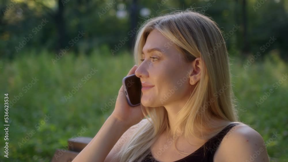 Attractive young woman talking on phone in park. blonde in black sports top sits on bench and chats on smartphone. girl is glad to hear answer and smile. Attractive young woman talking on phone in park. blonde in black sports top sits on bench and chats on smartphone. girl is glad to hear answer and smile.