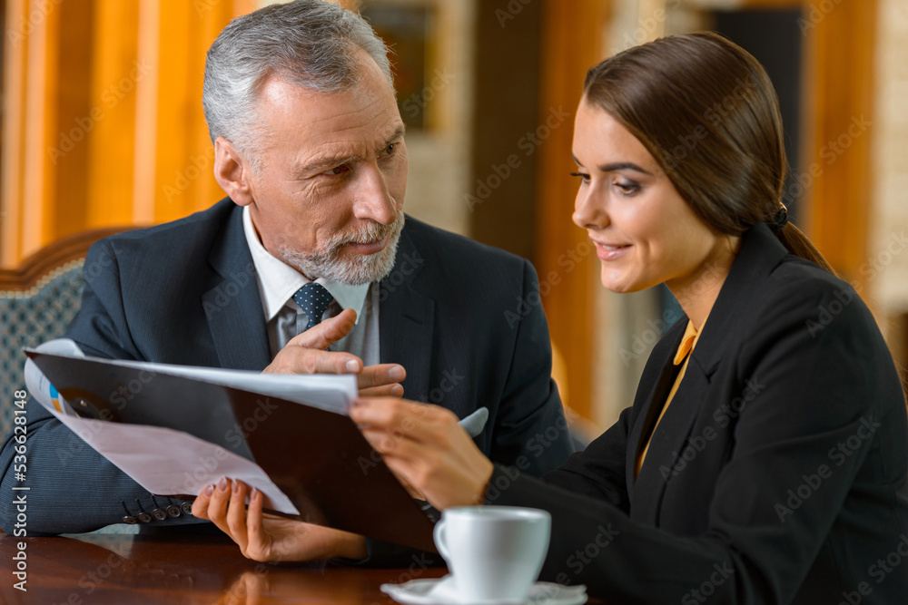 Young businesswoman showing documents to adult businessman while drinking coffee in luxury hotel
