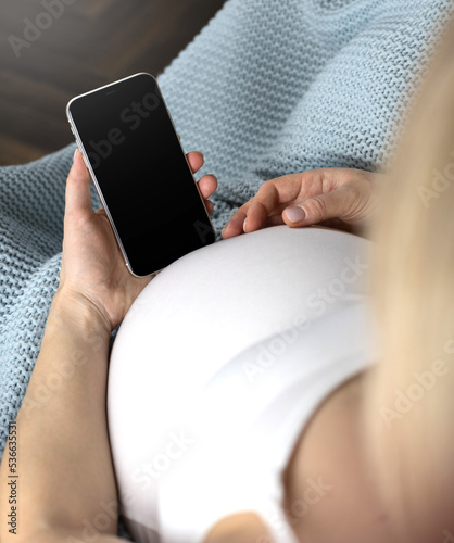 A pregnant woman is holding a telephone in her hands. A pregnant woman sits working at home covered with a blue blanket. woman looking at phone screen. High quality photo
