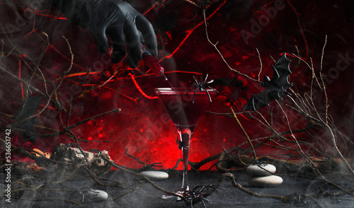 Halloween alcoholic cocktail bloody martini and hand with syringe on scary dark red background with twisted branches, bats, stones, pumpkin guards and spiders, festive drink for vampire party