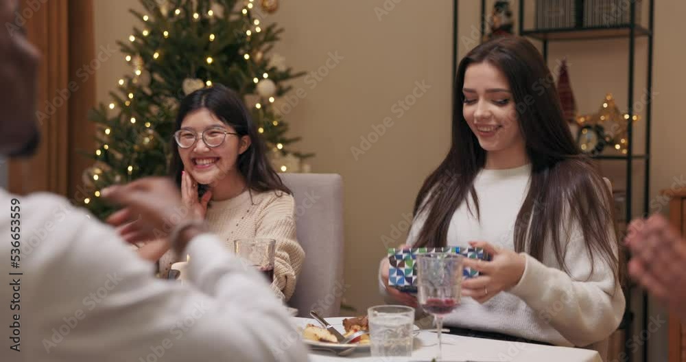 During party, guy gives gift to girlfriend. Brunette in white sweater unties the ribbon and opens the box.. She shows it to friends, thanks boyfriend and blows air kiss. Girl is pleasantly surprised. During party, guy gives gift to girlfriend. Brunette in white sweater unties the ribbon and opens the box.. She shows it to friends, thanks boyfriend and blows air kiss. Girl is pleasantly surprised.