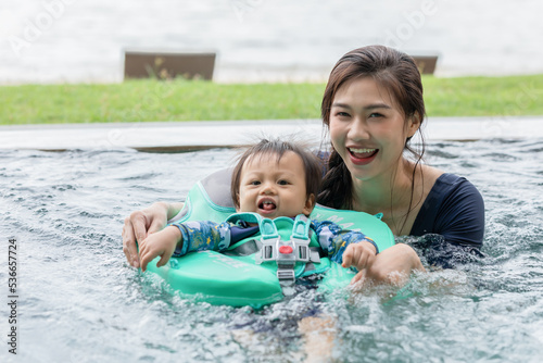 Happy Asian mom and baby playing in swimming pool using swim ring for leaning to swim relax in holiday. Vacation of family mother and baby spending time together with outdoor activity in swimming pool
