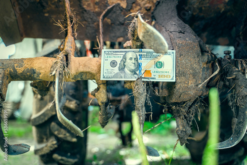 Hundred dollar bill between the teeth of a tractor rototiller close-up. Finance and agricultural machinery. Money on the immobilized tractor units. Stopping the work of the farm photo