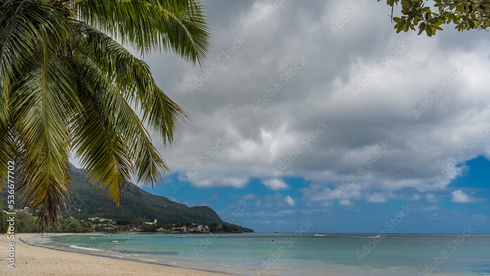 Beautiful sandy tropical beach. Boats are visible in the turquoise ocean. Hotel houses on a hill near the shore. Palm leaves against the sky and clouds. Seychelles. Mahe. Beau Vallon