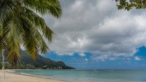 Beautiful sandy tropical beach. Boats are visible in the turquoise ocean. Hotel houses on a hill near the shore. Palm leaves against the sky and clouds. Seychelles. Mahe. Beau Vallon © Вера 