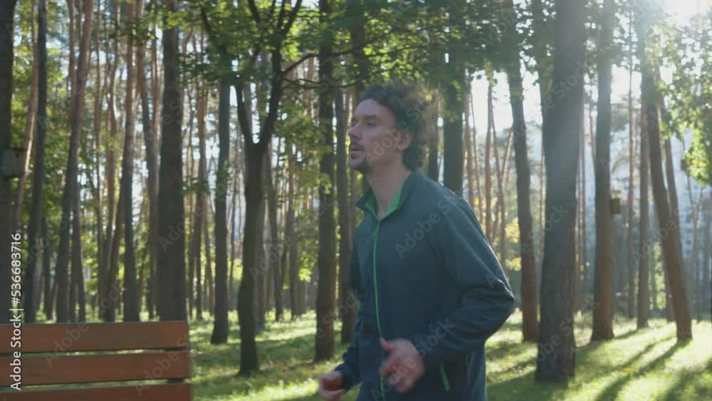 Slow motion young man with curly hair wearing sport jacket running in nature on sunny morning. Tracking shot of athlete against green trees. Healthy lifestyle. Concept of fitness Slow motion young man with curly hair wearing sport jacket running in nature on sunny morning. Tracking shot of athlete against green trees. Healthy lifestyle. Concept of fitness