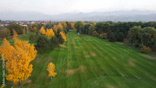 People play golf on a green field in autumn. Yellow-red leaves on some trees. Places for holes. There is a golf car. Houses and mountains are visible in the distance in a haze. Sunset. Wedding