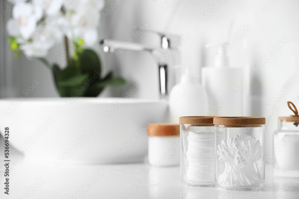 Glass jars with cotton pads and swabs on white countertop in bathroom. Space for text Glass jars with cotton pads and swabs on white countertop in bathroom. Space for text