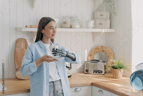 Young disabled girl holding mug by bionic prosthetic arm, enjoying domestic routine in cozy kitchen photo