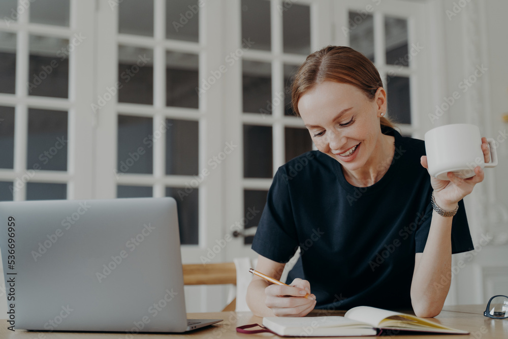 Smiling girl writing notes, planning schedule for day, holding coffee mug on workplace with laptop Smiling girl writing notes, planning schedule for day, holding coffee mug on workplace with laptop