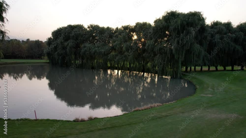 A green golf course with a view of a willow tree and a pond at sunset. The water in the pond is gray, reflecting the trees and sun. Birds are swimming. The sun is in a haze. Yellow-red leaves. Autumn A green golf course with a view of a willow tree and a pond at sunset. The water in the pond is gray, reflecting the trees and sun. Birds are swimming. The sun is in a haze. Yellow-red leaves. Autumn