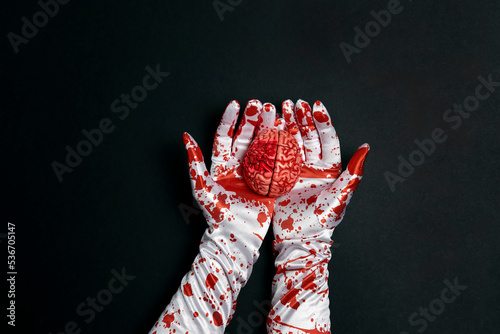 Creepy human brain in hands in bloody gloves on a black background. © WindyNight