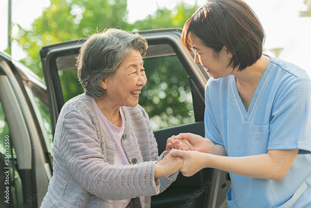 © One - シニア女性が車から降りるのを手伝う女性ヘルパー