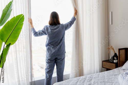 Young woman in pajamas open white curtains at the window, the morning after waking up in the bedroom of her home