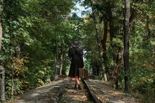Woman in black coat and hat of 40s style with retro valise in hands walks along rails going through forest. Back view