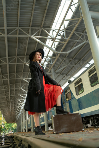Young woman on platform near train with leather suitcase wearing in red dress, black coat and hat . Vertical frame