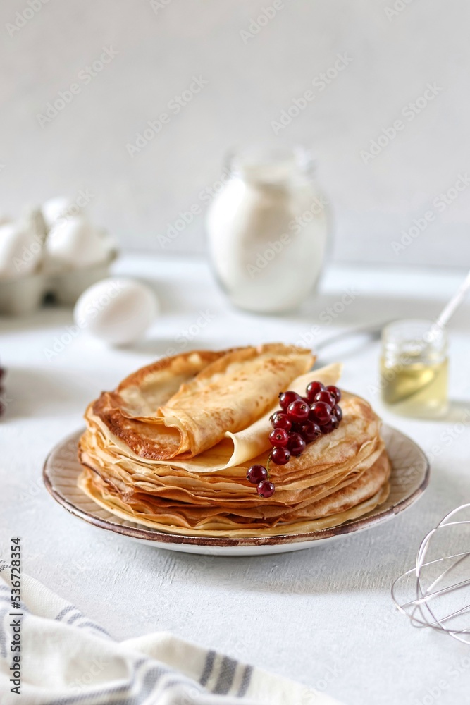 Thin pancakes in milk lie one on top of the other on a plate, currant berries lie on top. Nearby are eggs, flour and honey. Maslenitsa. Traditional cuisine.