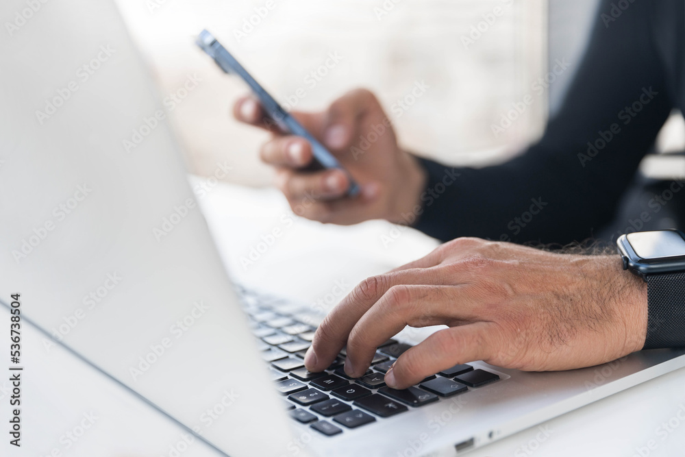 Fototapeta premium Man types on laptop keyboard and holds mobile phone, close-up photos